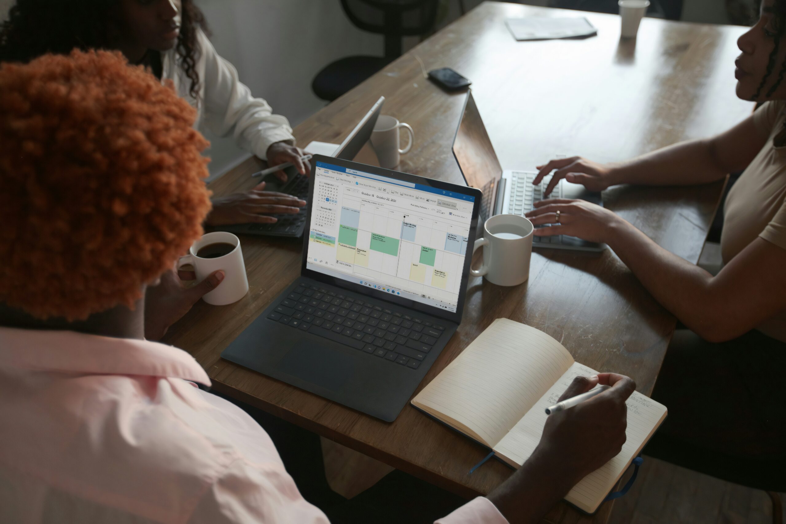 Introducing-the-New-Microsoft-Planner-Everything-You-Need-to-Know–scaled.jpg Three people in a meeting at a table discussing schedule on their Microsoft laptop